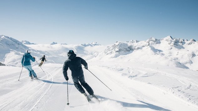 Drei Skifahrer auf einer schneebedeckten Piste mit Bergen im Hintergrund unter klarem Himmel.