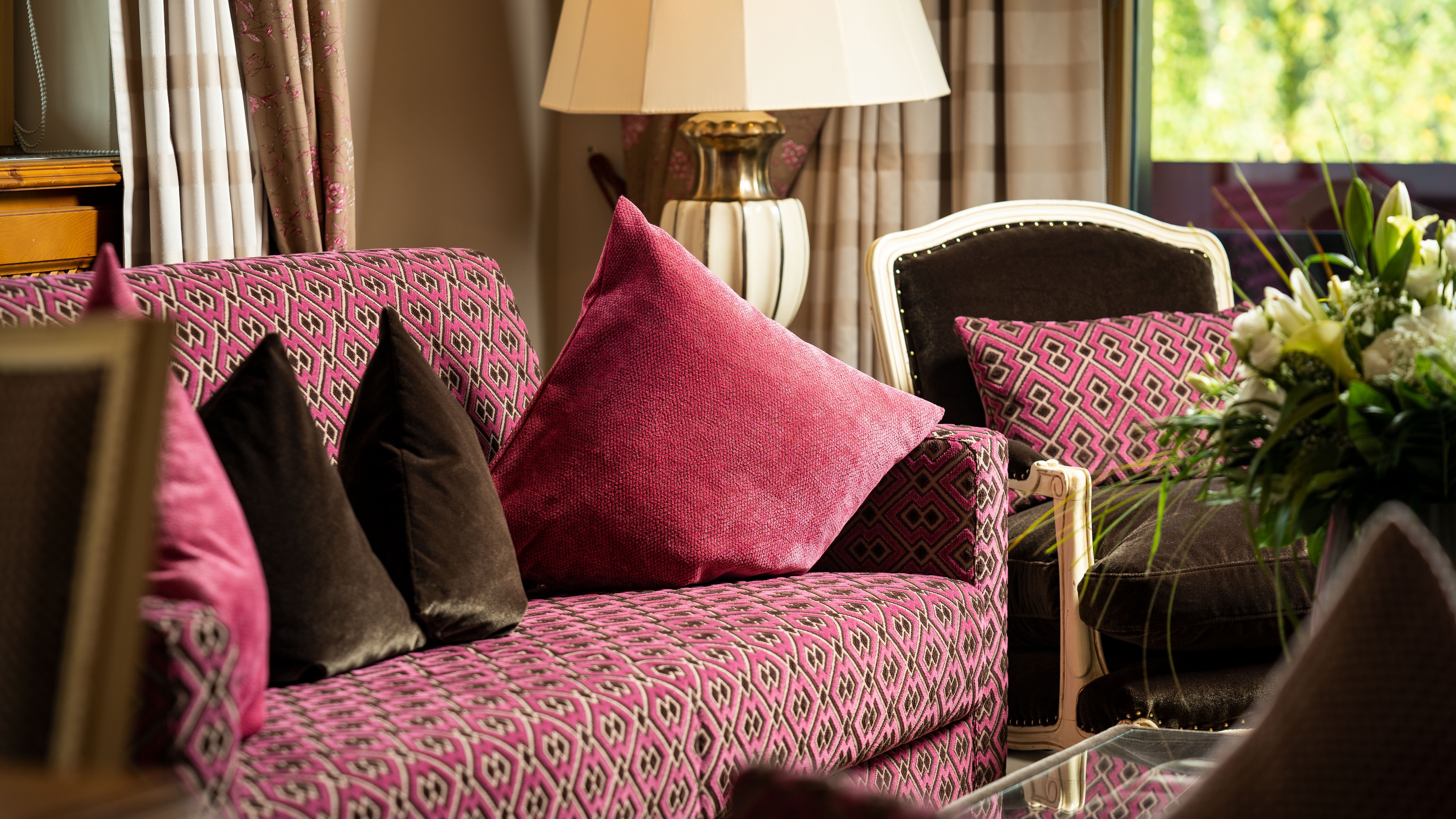 Romantik Suite Ein stilvolles Wohnzimmer mit einem gemusterten rosa Sofa, dekorativen Kissen, einer Lampe und Pflanzenarrangement.