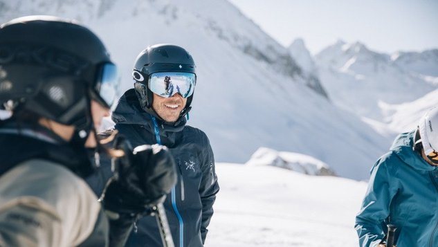 Gruppe von Skifahrern auf einer verschneiten Piste, ein Skifahrer trägt Helm und Brille, lächelt