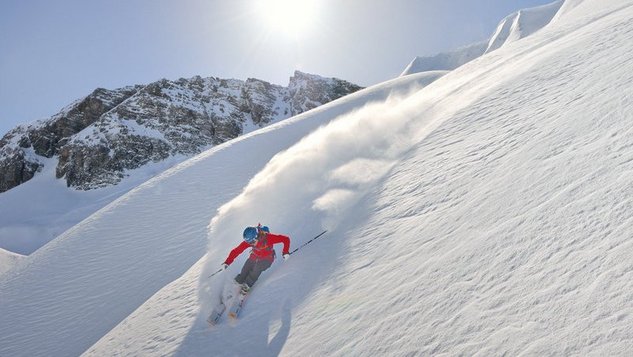 Skifahrer auf schneebedecktem Hang mit Bergen im Hintergrund unter hellem Sonnenlicht