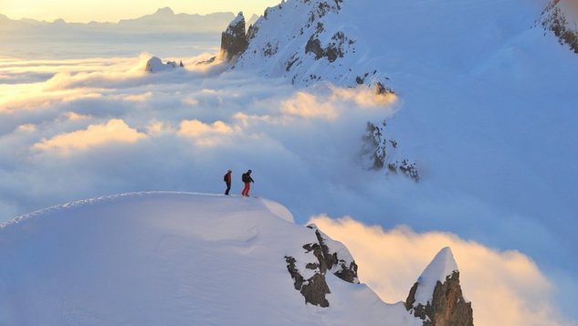 Zwei Personen stehen auf einem schneebedeckten Berggipfel umgeben von Wolken und einer Berglandschaft im Hintergrund.
